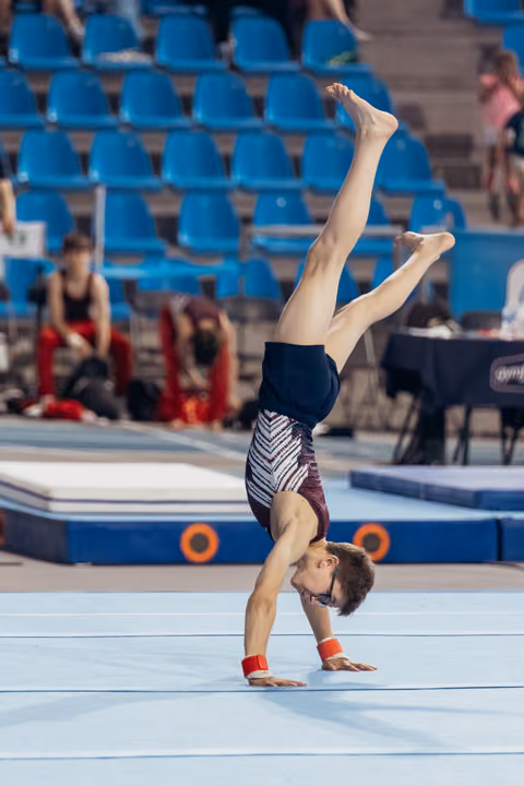 Young male gymnast performs a handstand with legs split on the floor exercise mat, wearing red wristbands.