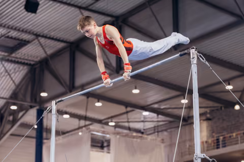 Young male gymnast grips the high bar with intense focus, body horizontal, performing in an indoor sports hall.
