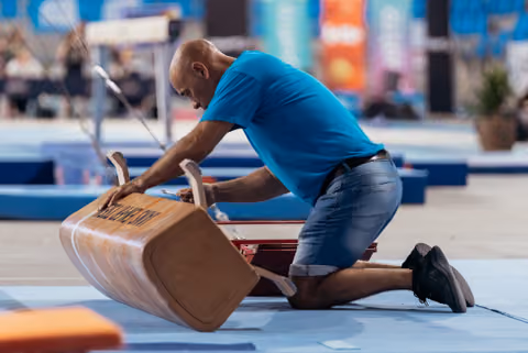 Focused bald man in blue shirt kneels on mat, gripping a tipping wooden vaulting box with intense concentration.