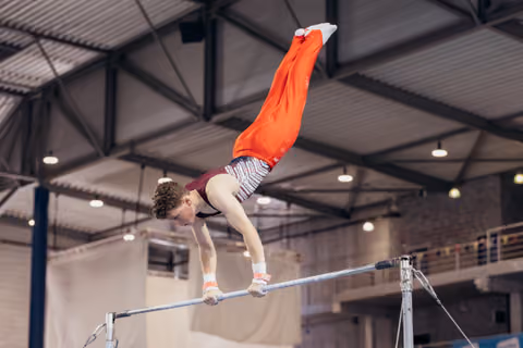 Young male gymnast performs an inverted handstand on the high bar, legs extended upward in red pants, inside a sports hall.