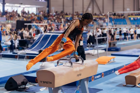 Young gymnast in orange performs on the pommel horse, leaning forward with intense focus during a busy competition.