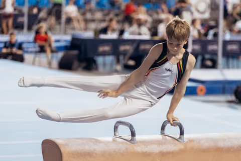 Young male gymnast performs a focused pommel horse routine, legs extended, intense concentration on his face at a gymnastics meet.