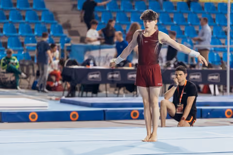 Young male gymnast stands focused on the floor exercise mat, arms extended, barefoot, in a maroon leotard during a competition.