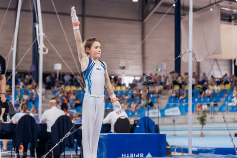 Young male gymnast raises hand to salute judges, standing confidently on podium in white and blue Agiva leotard.