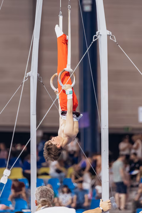 Young male gymnast performs inverted hang on still rings, shirtless in red pants, intense concentration in packed arena.