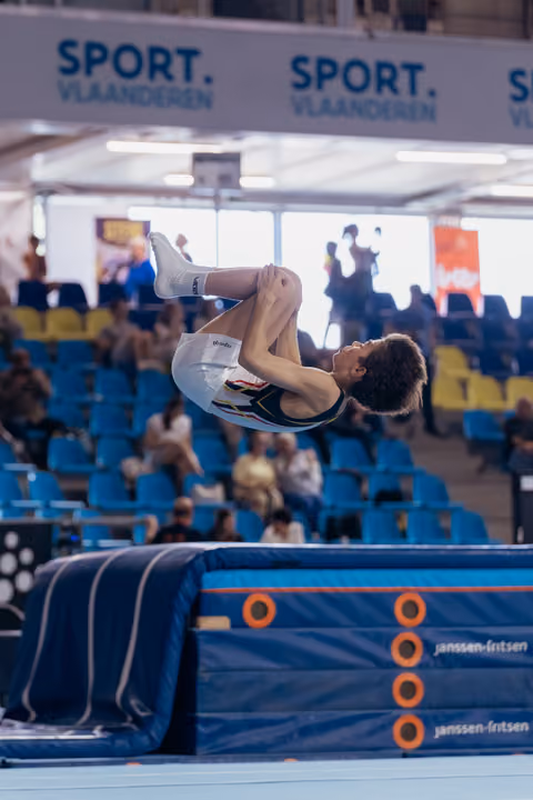 Young male gymnast performs a tight tuck backflip above the trampoline bed, focused and airborne in a packed arena.