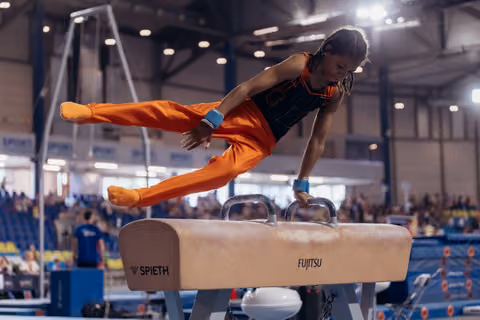 Young gymnast in orange performs a powerful split on the pommel horse at an indoor competition, focused and intense.