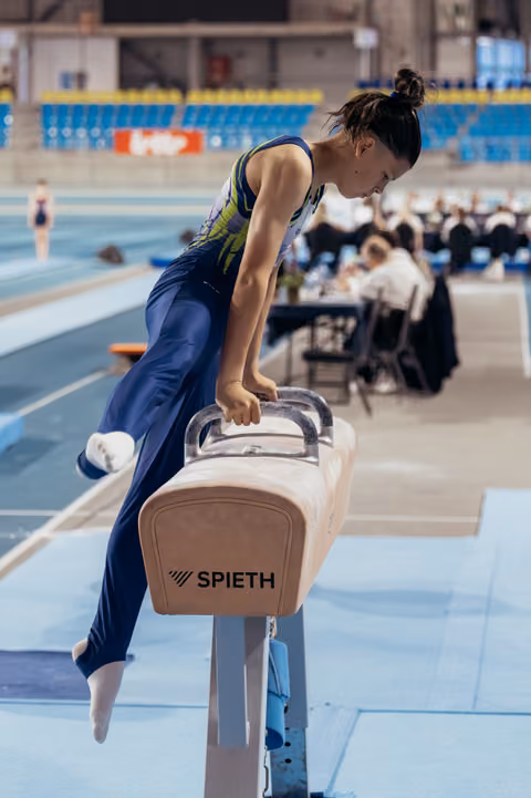 Young female gymnast concentrates intensely while performing on a Spieth pommel horse in a large indoor arena.