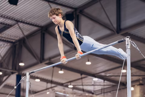 Young male gymnast grips the high bar with concentration, body extended horizontally in an indoor gymnasium.