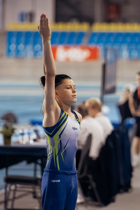 Young gymnast in blue and green leotard raises hand to salute judges, eyes closed, composed expression in arena setting.