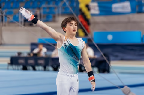 Young male gymnast raises arm in salute, wearing teal leotard and grips, focused expression on blue arena floor.