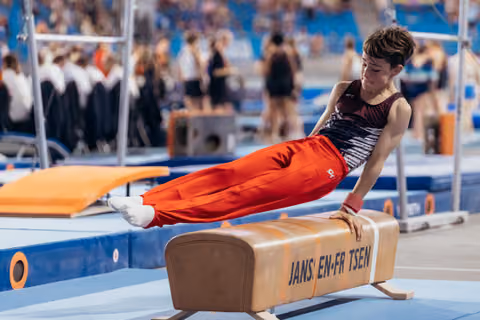 Young male gymnast performs pommel horse routine, body parallel to floor, focused expression, in a competition arena.