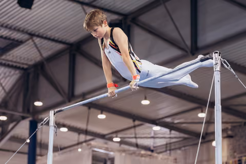 Young male gymnast grips the high bar with intense focus, legs extended mid-routine in an indoor gymnasium.