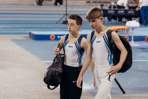 Two young male gymnasts in Agiva leotards walk purposefully through the arena, bags in hand, expressions serious and focused.