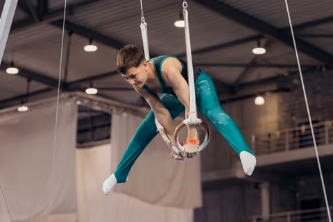 Young male gymnast grips still rings mid-routine, body coiled with intense focus, in a large indoor gymnasium.