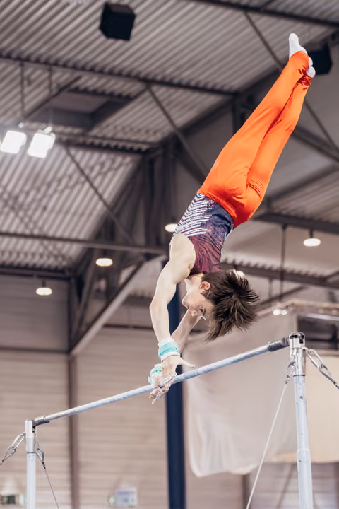 Young male gymnast performs a handstand on the horizontal bar, body fully extended upward in orange competition leotard.