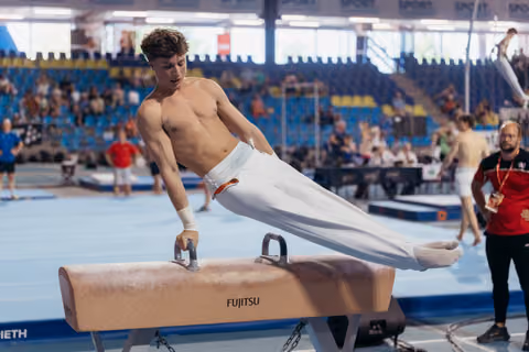 Young male gymnast performs on a Fujitsu pommel horse, body horizontal, expression focused during a competition.