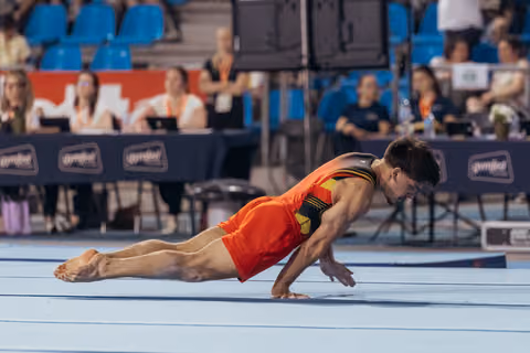 Male gymnast in red uniform executes a dramatic floor dive, body parallel to the mat at a gymnastics competition.
