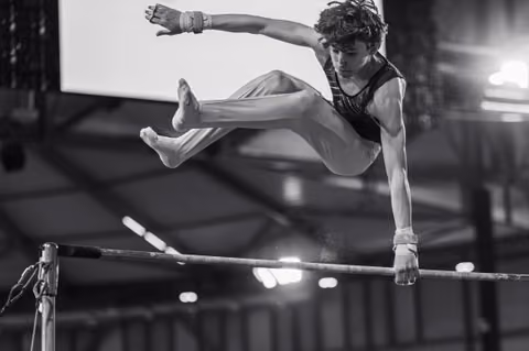 Young male gymnast mid-flight above the high bar, legs split, intense focus, wearing wrist guards in an indoor arena.