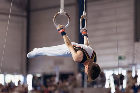 Male gymnast performs an inverted iron cross on still rings, body horizontal, intense focus during competition.