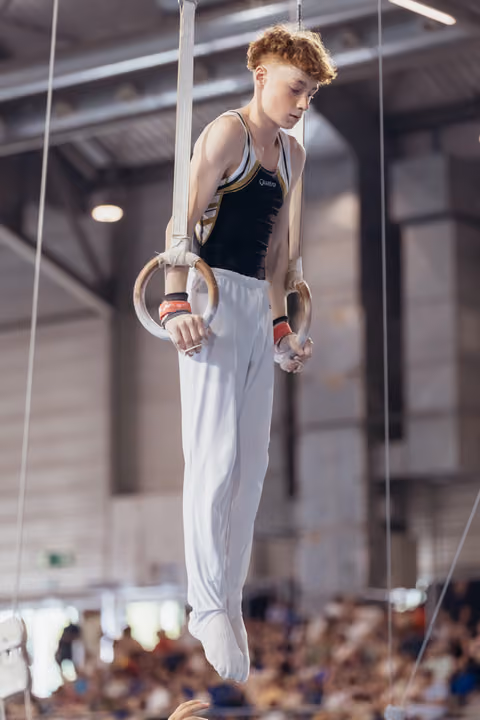 Young male gymnast with red hair grips still rings, looking down with intense focus during a gymnastics competition.
