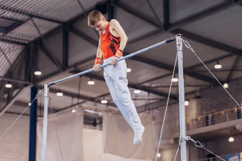 Young male gymnast grips the high bar with intense focus, legs extended mid-routine in an indoor gymnastics hall.