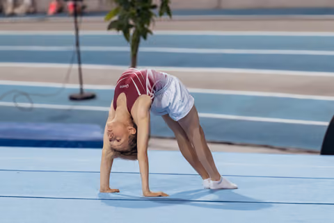 Young gymnast performs a backbend bridge on a blue gymnastics floor, focused and flexible during a sports event.