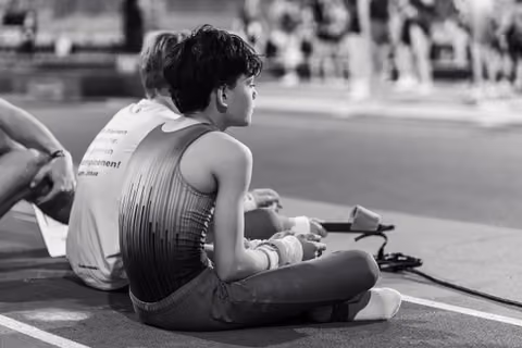 Young gymnast sits cross-legged on the floor, wrists wrapped, watching intently with quiet focus during a gymnastics event.