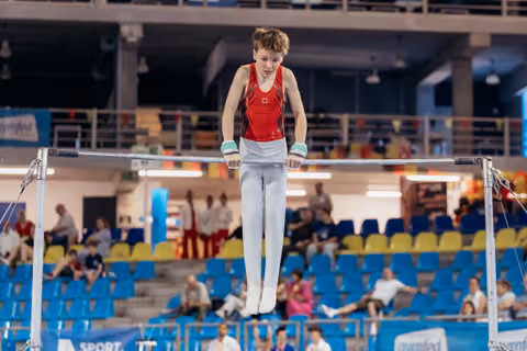 Young male gymnast grips the high bar with intense focus, body suspended mid-routine in a packed sports arena.