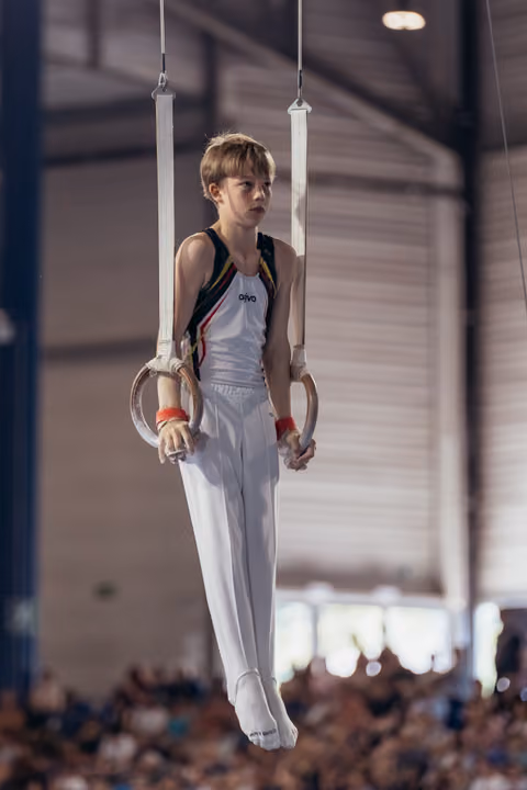 Young male gymnast hangs with focused expression on still rings during an indoor gymnastics competition.