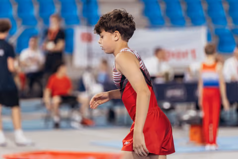 Young male gymnast in red leotard stands focused and composed, preparing for his routine in a busy sports arena.