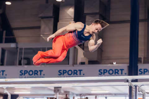 Male gymnast soars above parallel bars in red and navy uniform, body tucked, face intense with effort.