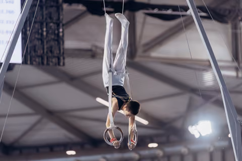 Young male gymnast holds an inverted cross position on still rings, face showing intense concentration in a competition hall.
