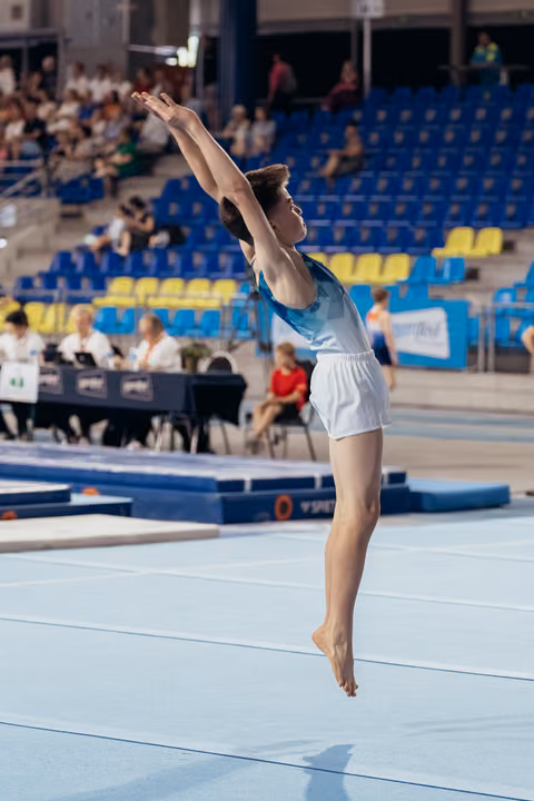 Young male gymnast mid-leap on floor exercise, arms reaching skyward, poised and focused during competition.