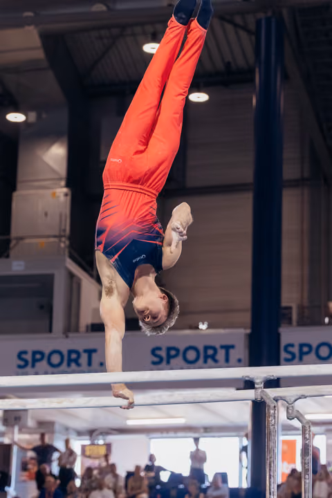 Male gymnast holds a perfect handstand on parallel bars, body fully extended, focused expression, at an indoor sports event.