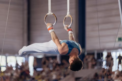 Young male gymnast performs an inverted hold on still rings, body fully extended, face taut with concentration.