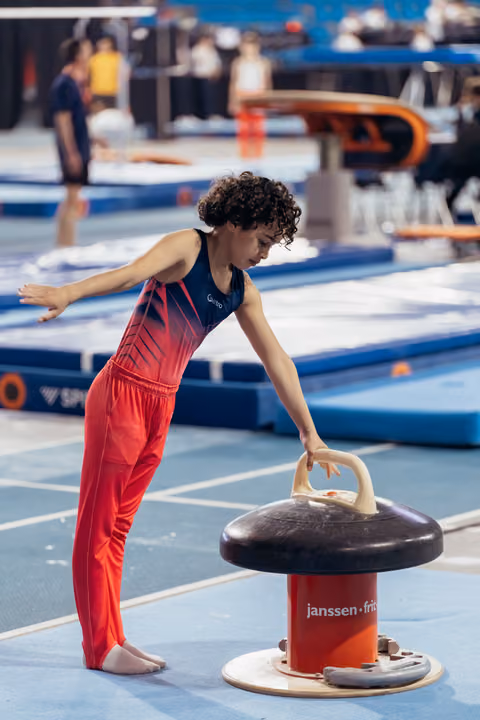 Young female gymnast with curly hair leans over pommel horse, focused and deliberate, in a busy competition hall.