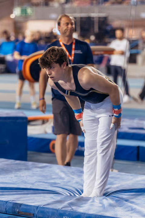 Young male gymnast lands on mat, leaning forward with intense focus, coach watching attentively in background.