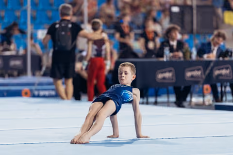 Young boy gymnast performs a floor routine, holding a low arabesque position with intense focus on his face.