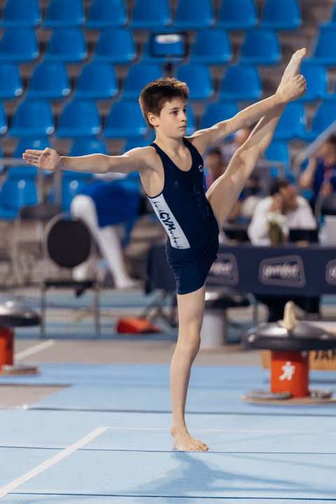 Young male gymnast performs a standing scale on the floor, arms extended, face calm and concentrated during competition.