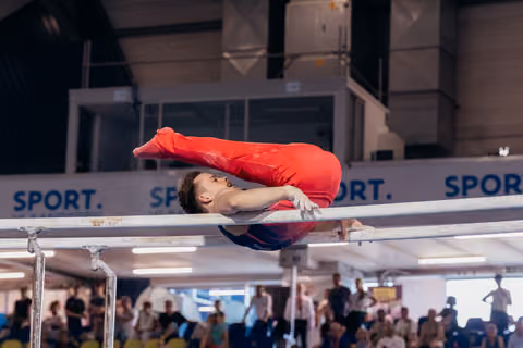 Male gymnast in red performs a tucked pike hold on parallel bars, body folded with focused expression, crowd watching.