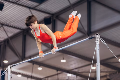 Young gymnast in red uniform grips the horizontal bar with intense focus, legs raised in a pike position indoors.