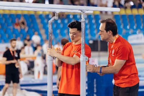 Two coaches in red shirts carefully adjust parallel bars at a gymnastics event, focused and deliberate in their setup.