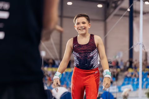 Young male gymnast beams with pride after his high bar routine, wearing red leo and wrist guards in a busy arena.