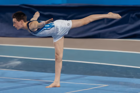 Young male gymnast holds a precise arabesque balance on floor, arms swept back, one leg extended high, focused expression.