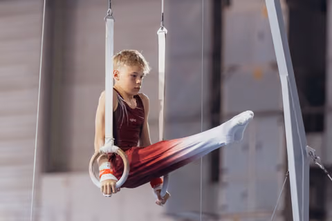 Focused young male gymnast holds a tuck position on still rings, wearing red and white competition attire.