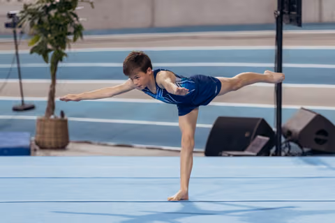 Young male gymnast holds a precise arabesque balance on floor, arms extended, focused expression in a competition hall.