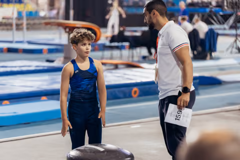 Young male gymnast listens attentively to coach's instructions before vault, both standing on competition floor.