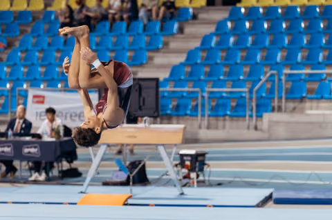 Young gymnast performs an inverted flip over the pommel horse, body fully extended, in a sparsely filled arena.