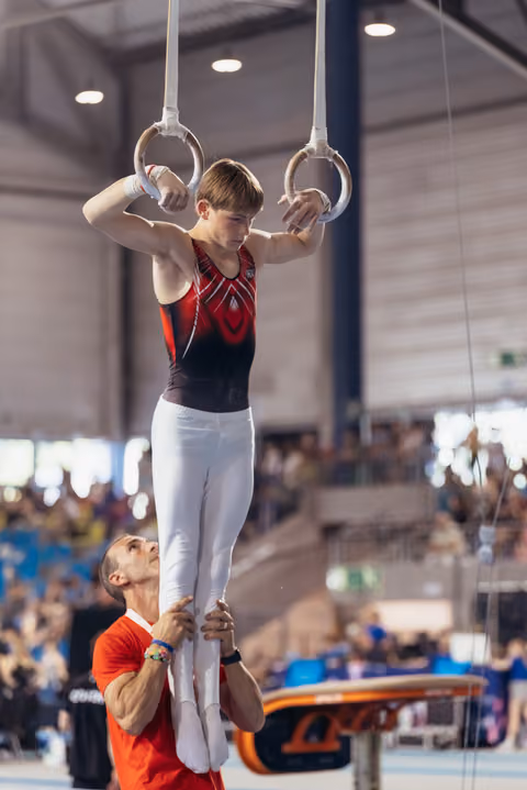 Young male gymnast grips still rings with intense focus while coach steadies his legs in a packed arena.